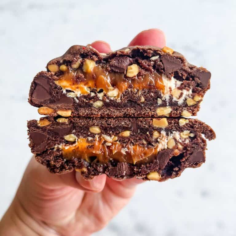 Thick chocolate cookies with a caramel and peanut filling being held against a marble background.